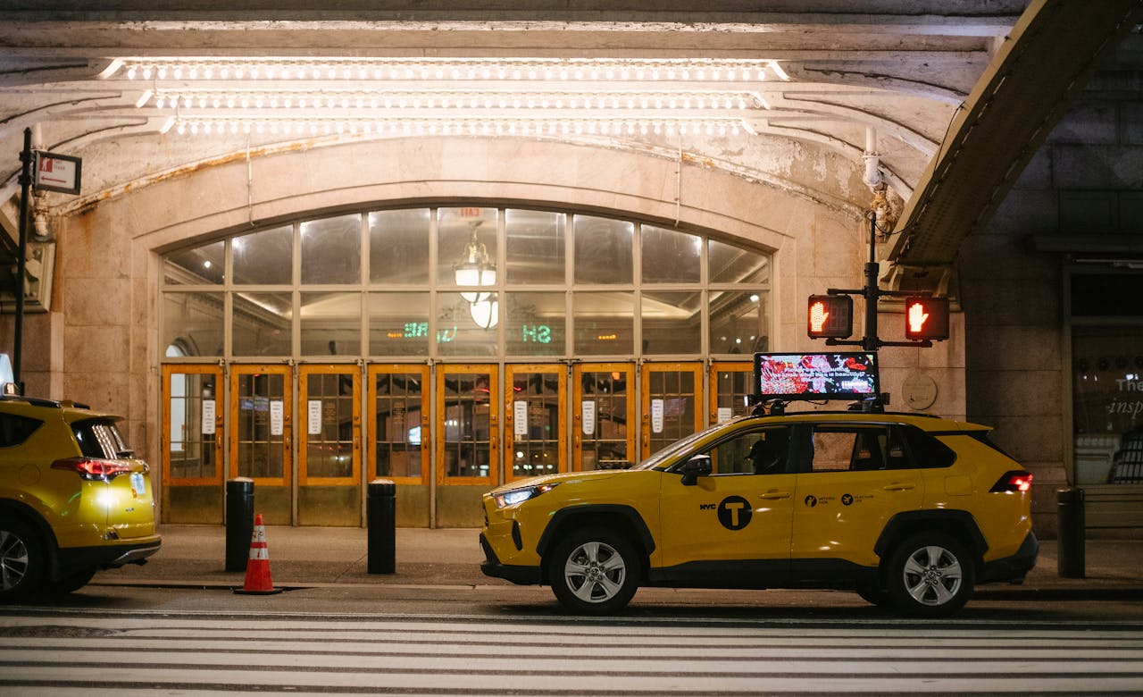 A yellow taxi cab parked outside a well-lit building entrance at night in an urban setting.